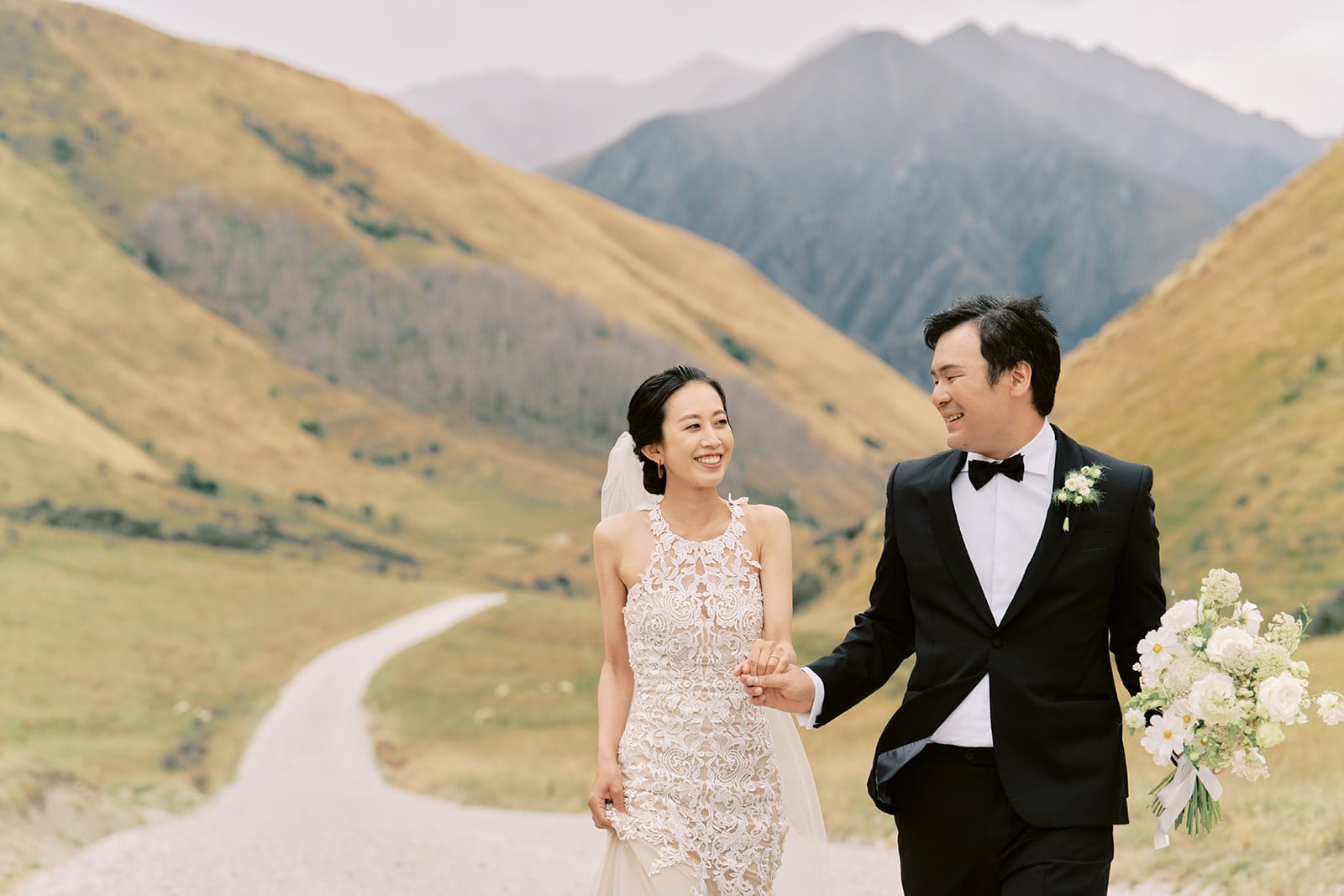 Queenstown New Zealand Heli Wedding Elopement Photographer クイーンズタウン　ニュージーランド　エロープメント 結婚式 | A couple in wedding attire smiling and holding hands while walking down a countryside road with mountains in the background, expertly captured by a Queenstown Wedding Photographer.